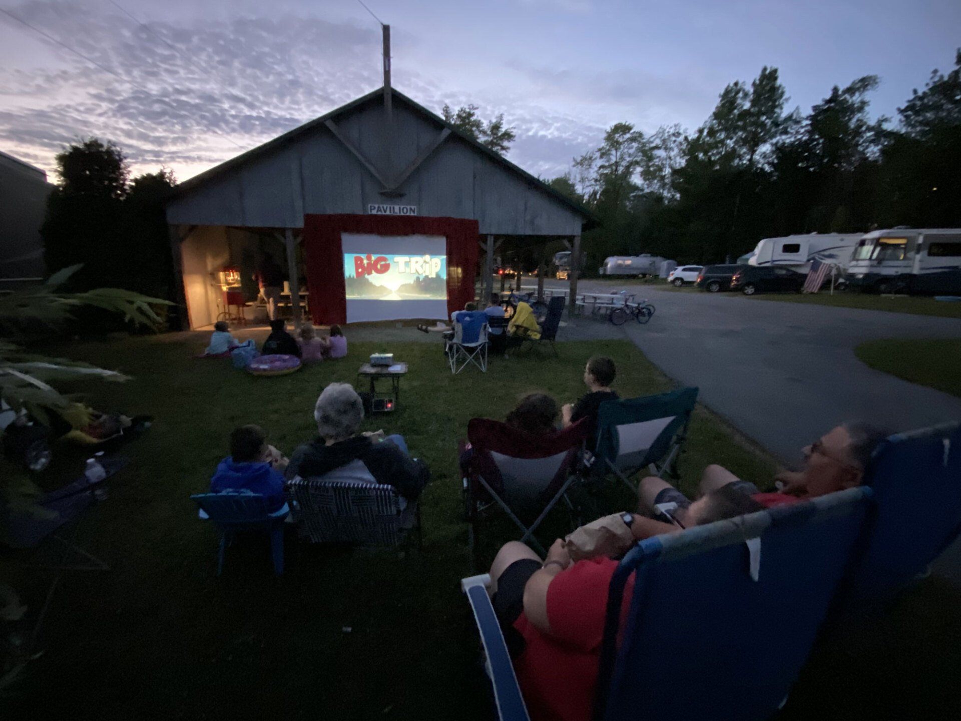 People seated outdoors watching a movie projected on a barn at dusk, with RVs parked nearby.