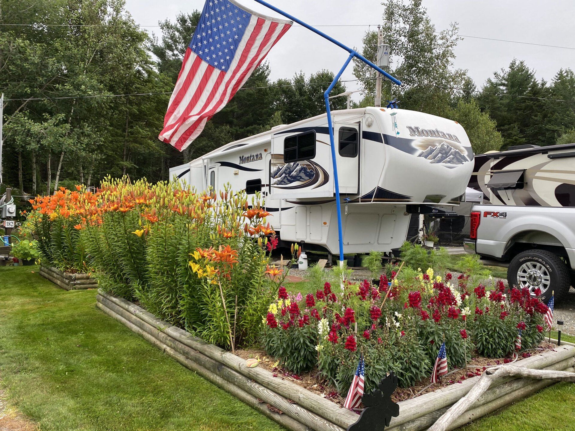 Camper trailer beside a flower bed with colorful flowers and a large American flag outdoors