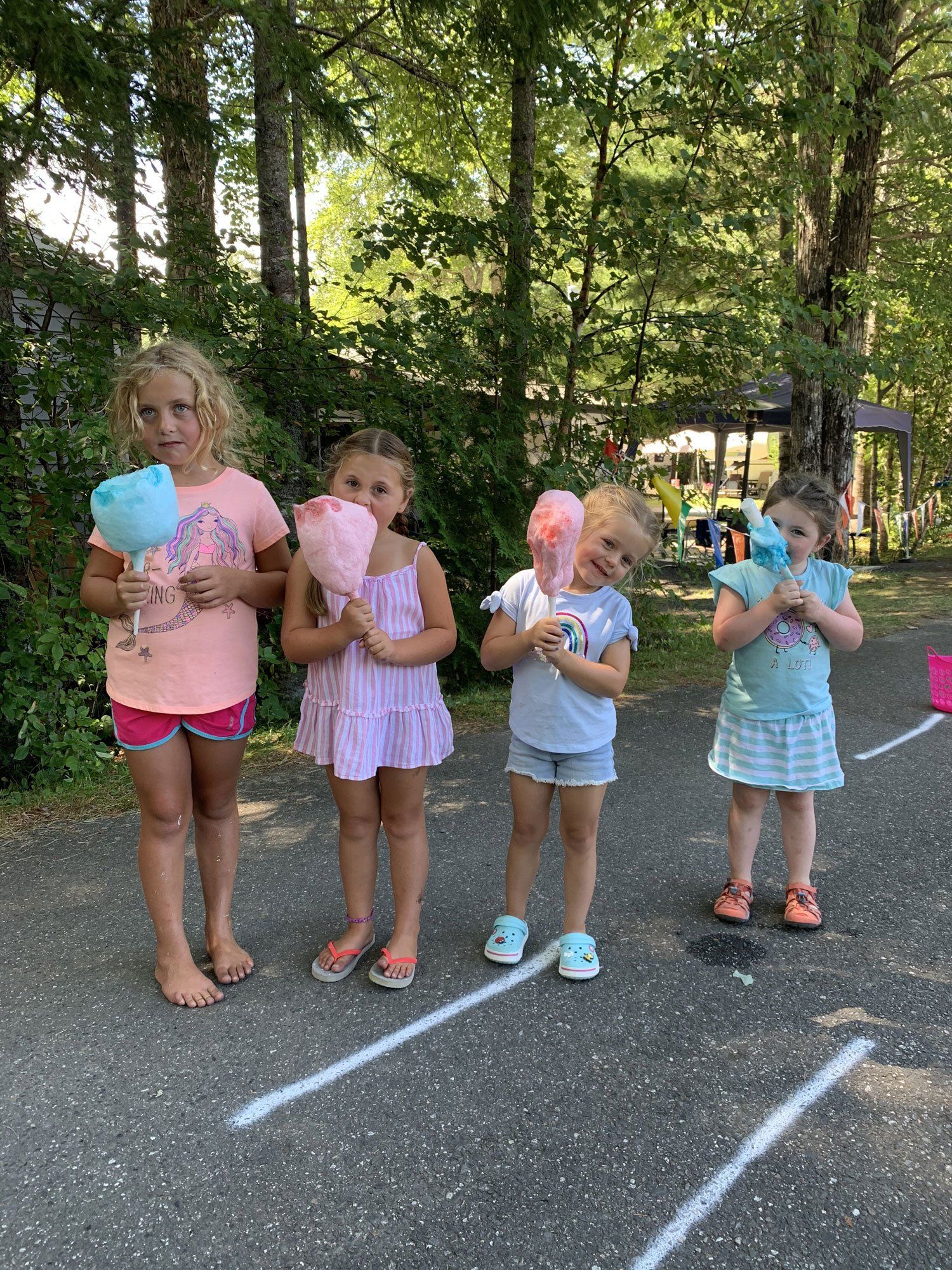 Four children outdoors holding cotton candy, standing on a paved path by trees.