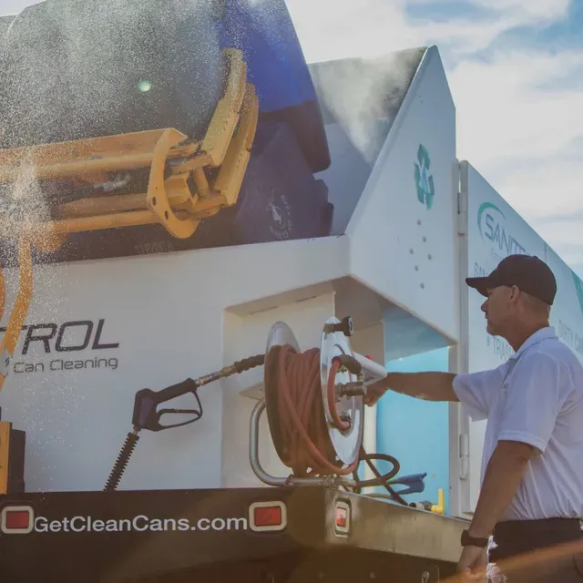 Man cleaning a recycling bin on a truck, white truck with water hose, sunny day.