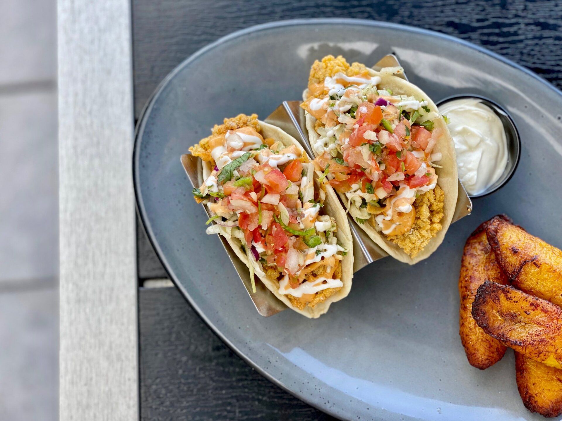 Two fish tacos on a blue plate with cream sauce and a side of fried plantains
