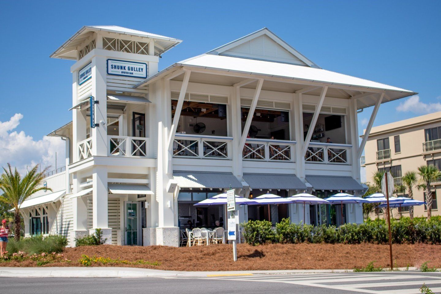 White beachfront restaurant with outdoor patio, balcony seating, and blue umbrellas under a bright sky