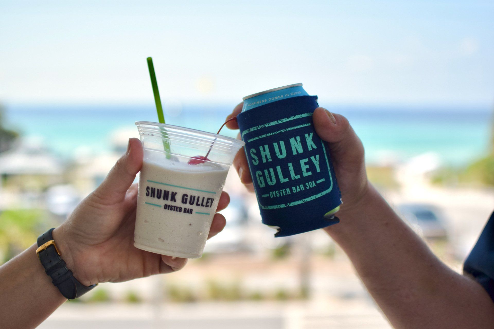 Two hands clink beach drinks with ocean view in the background