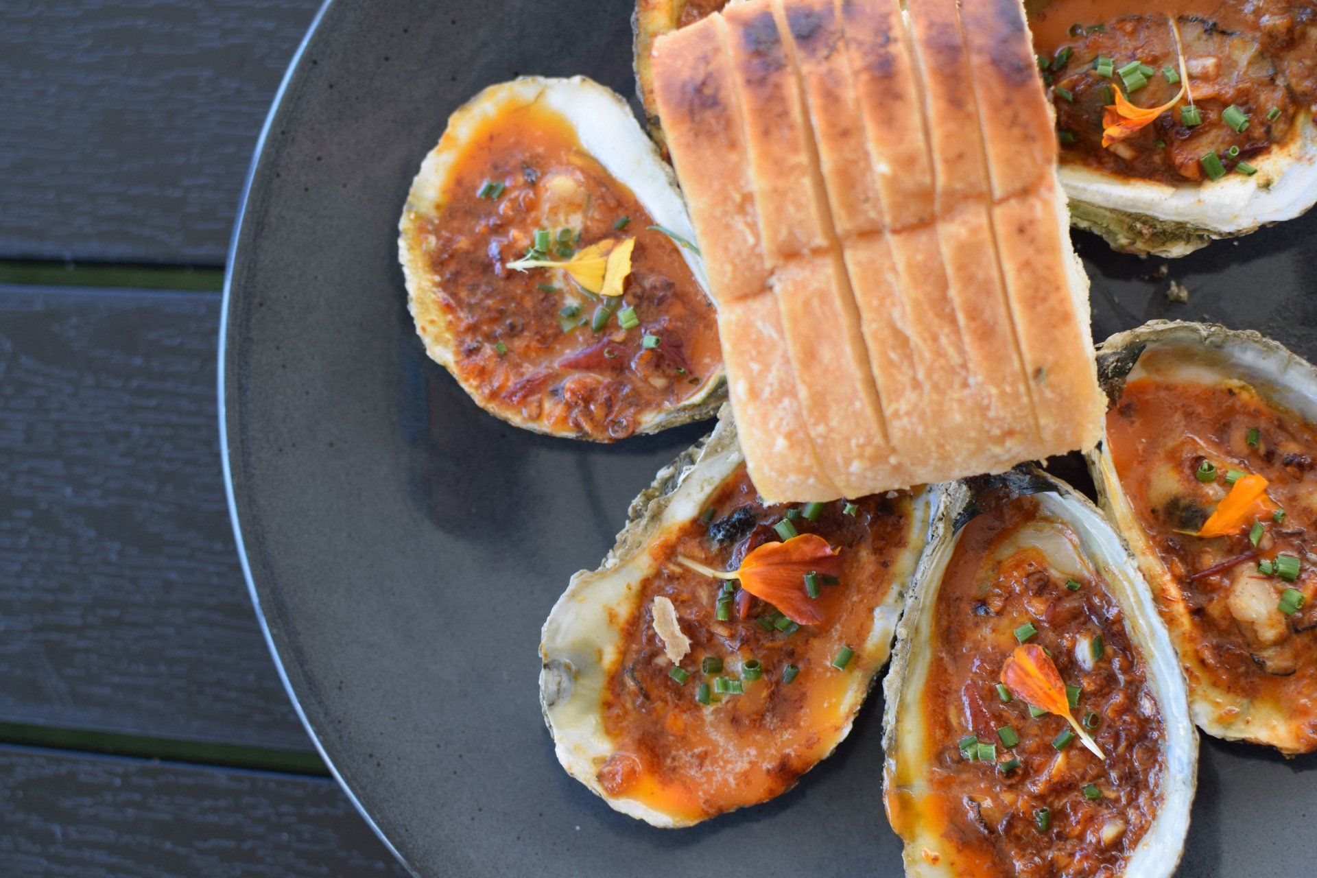 Grilled oysters topped with herbs on a dark plate, with a piece of bread on top