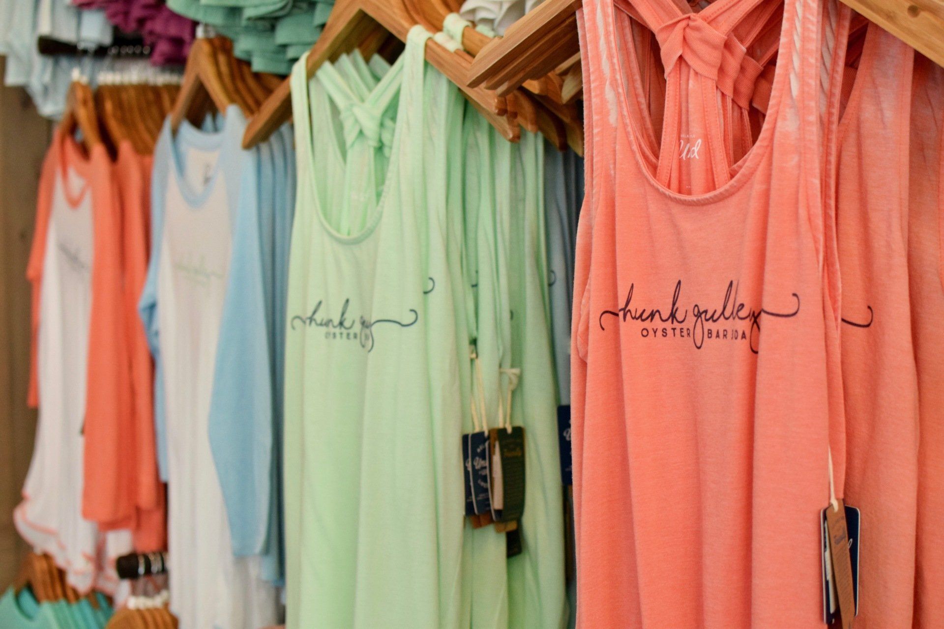 Colorful sleeveless shirts on hangers in a clothing store, including coral, mint, and blue tops.