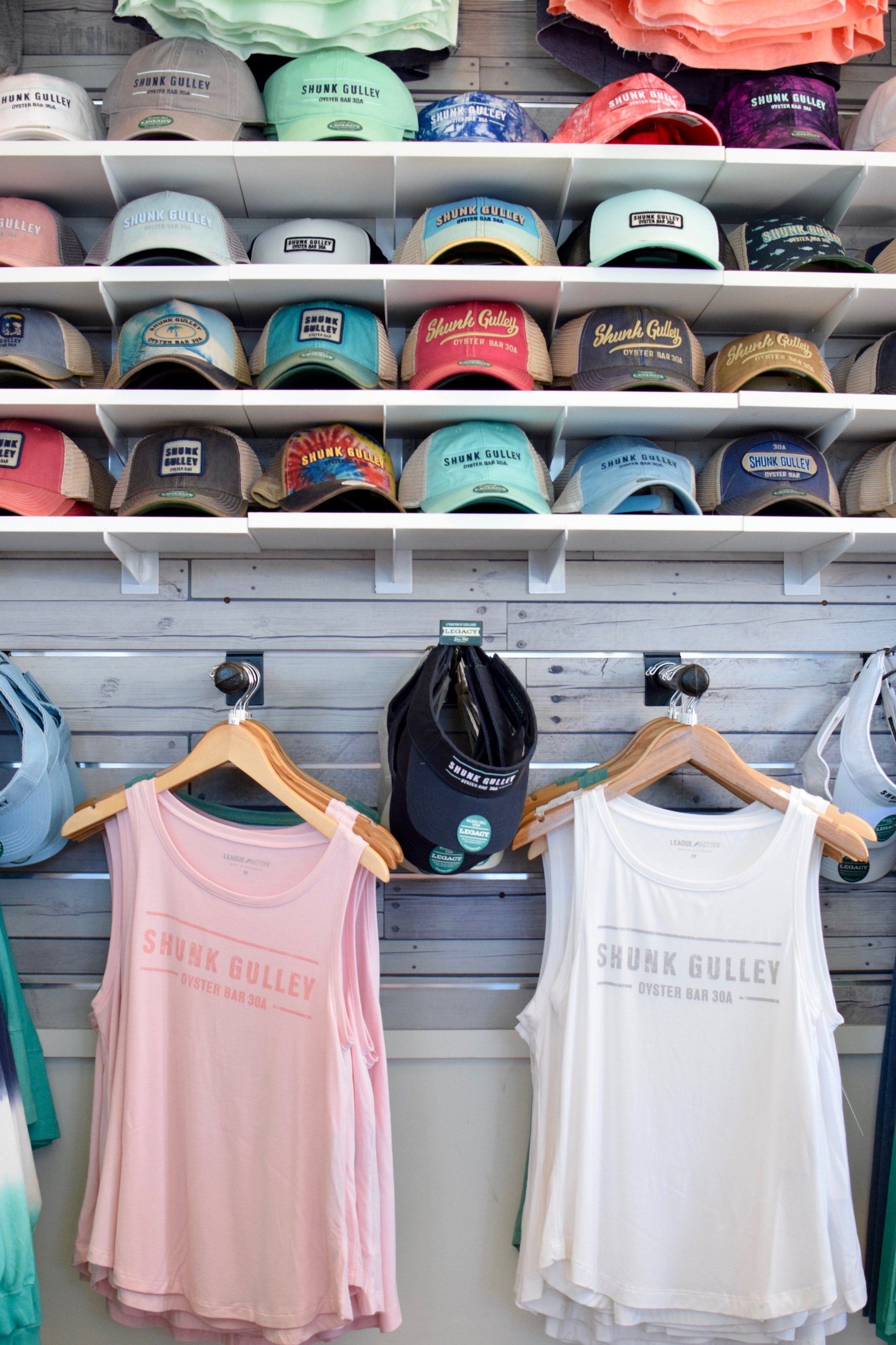 Hat display on shelves above hanging tank tops in a clothing store, with pink and white shirts below.