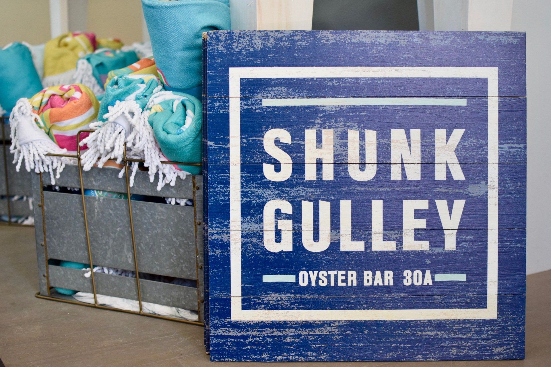 Blue sign reading “SHUNK GULLEY OYSTER BAR & GRILL” beside a crate of colorful beach toys