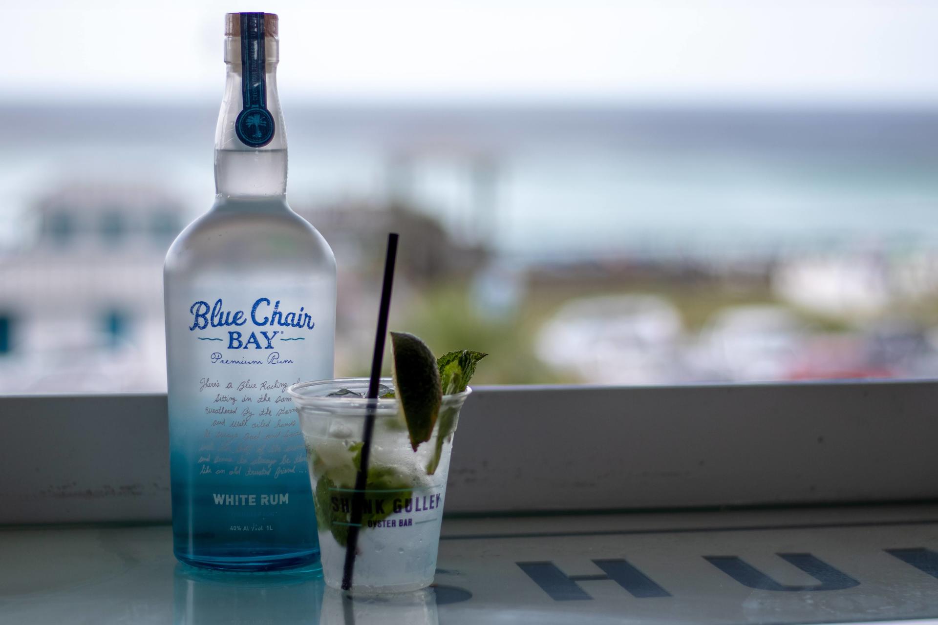 Blue Chair Bay bottle beside a lime cocktail on a window ledge, with a blurred ocean view in the background