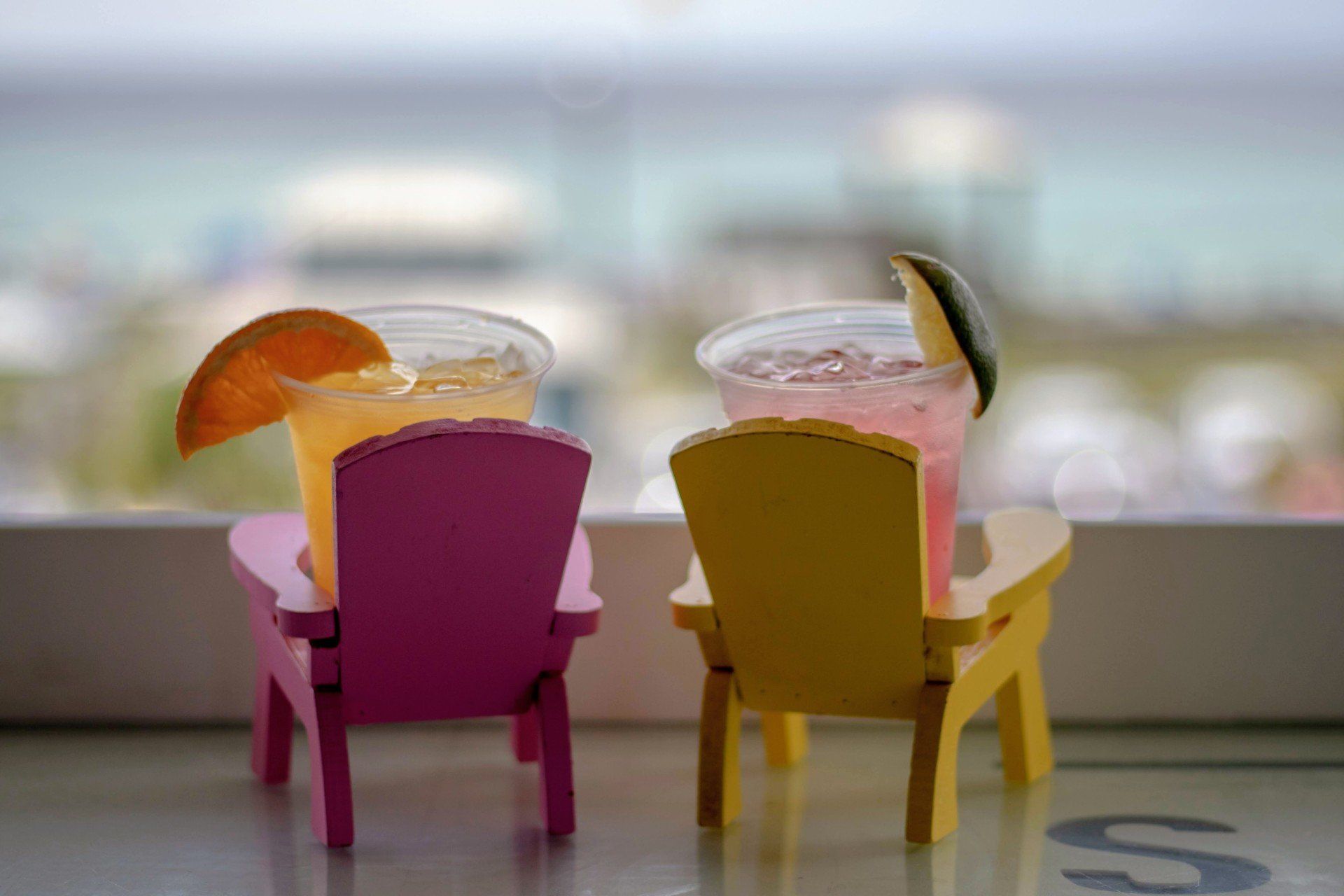 Two colorful tropical drinks in small chairs on a table, with blurred beach in background
