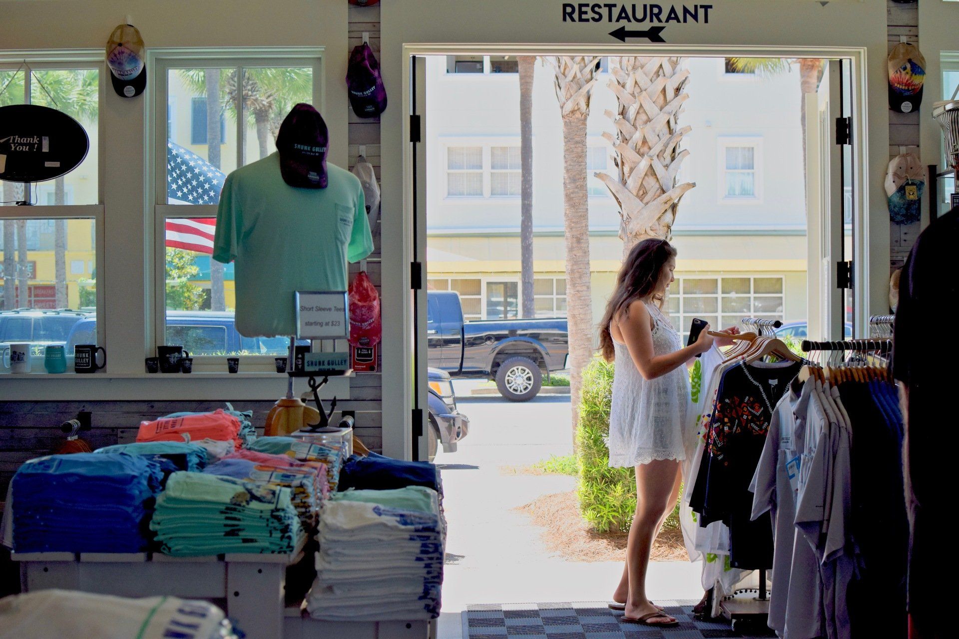 Clothing shop interior with folded shirts, a mannequin, and a woman browsing racks by the entrance