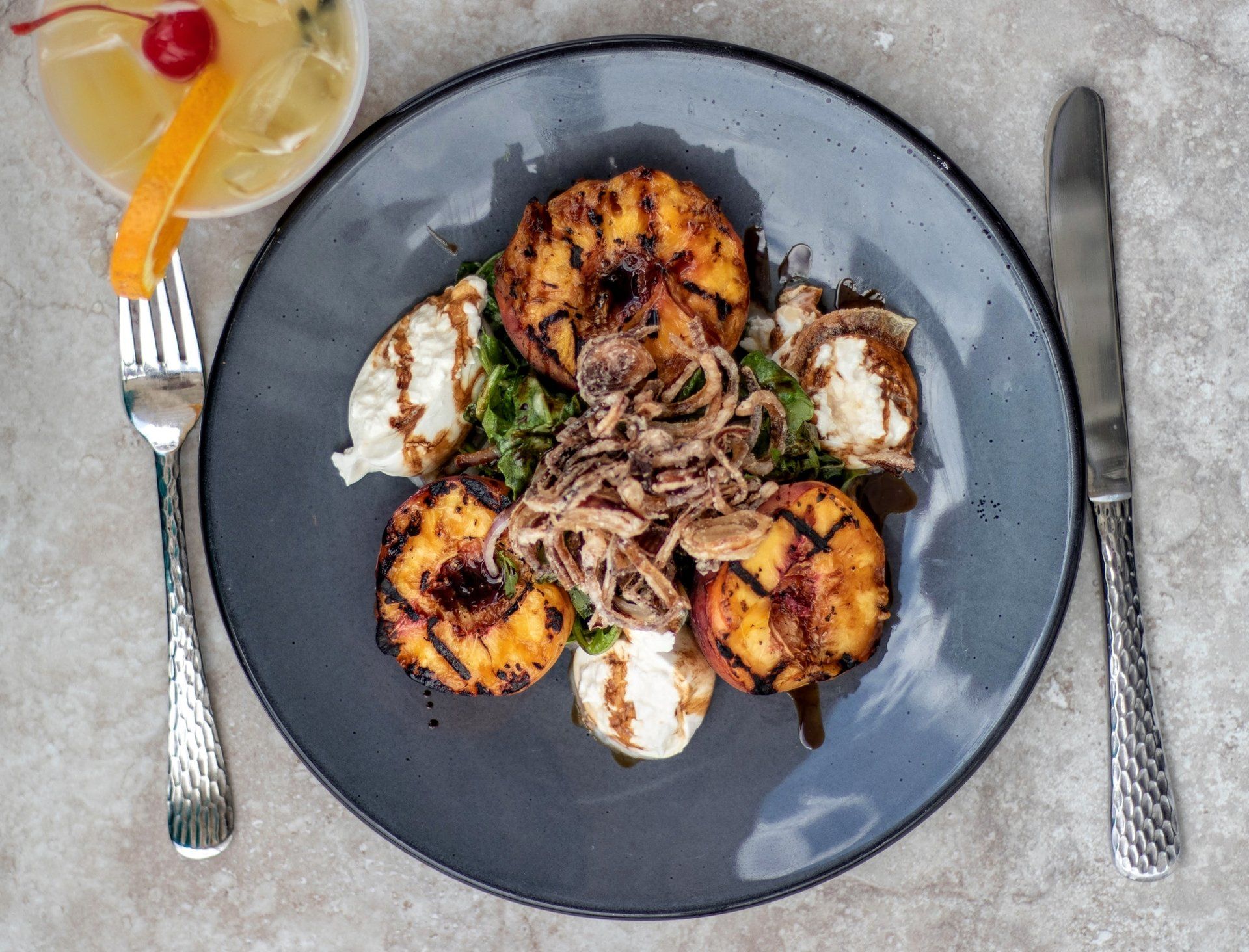 Grilled peaches and whipped cheese on a blue plate with nuts, served beside a fork, knife, and a cocktail.