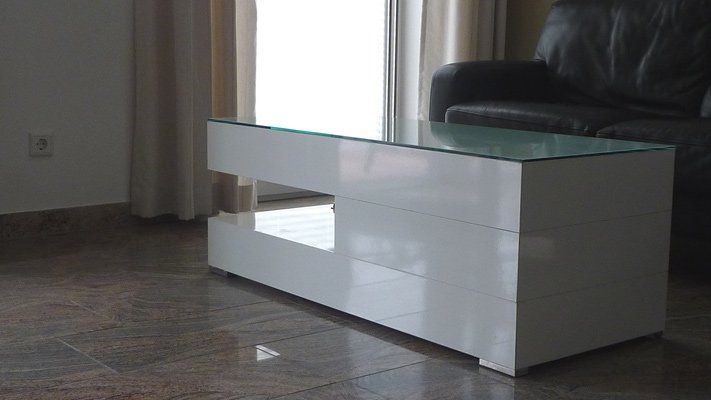 Ein weißer Couchtisch mit Glasplatte steht in einem Wohnzimmer neben einem Sofa.