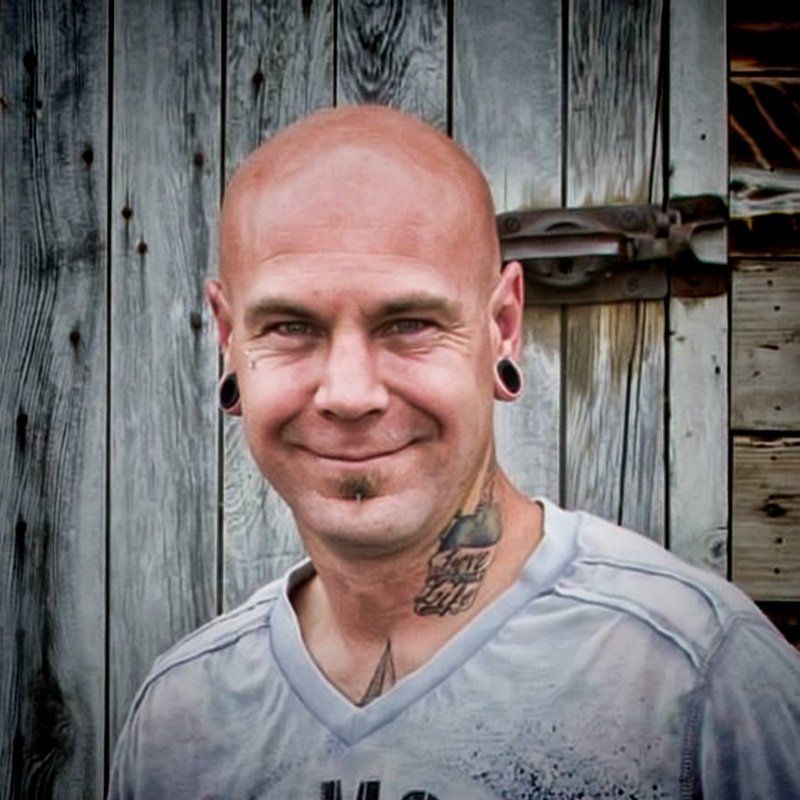 A bald man with a tattoo on his neck is smiling in front of a wooden wall.