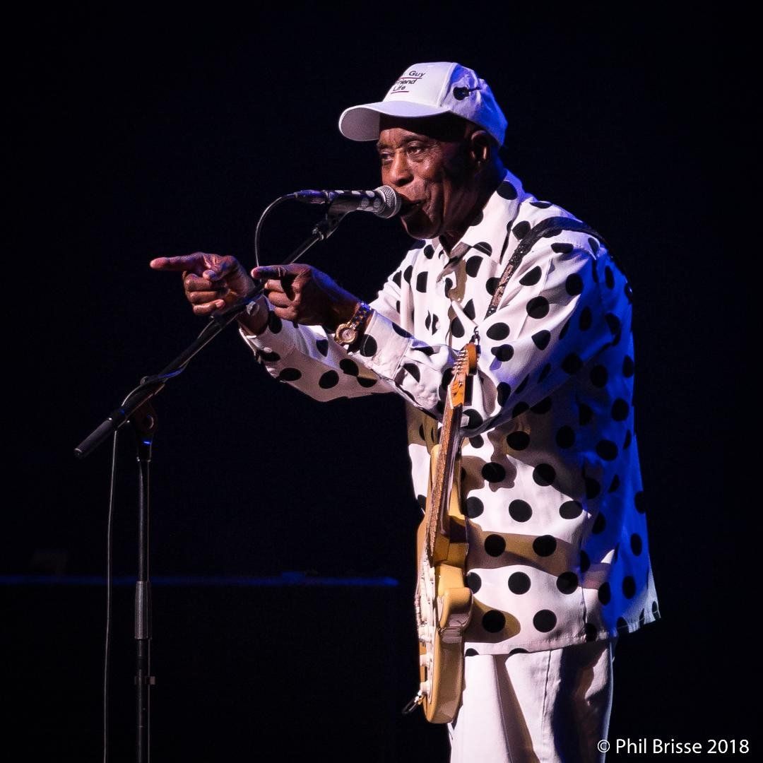 Buddy Guy on stage, pointing both fingers at the audience and smiling