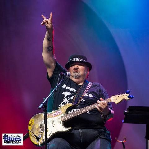 Popa Chubby sur les planches, assis avec sa guitare avec son bras dans les airs