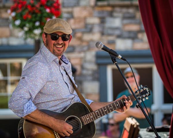 Steve Strongman playing guitar and grinning widely at the photographer wearing sun glasses on a bright sunny day at the Deslauriers Stage in Tremblant.