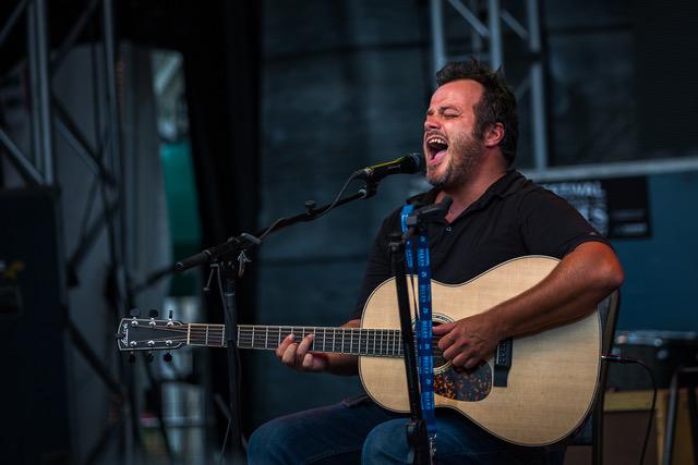Adam Karch singing and playing guitar sitting down.