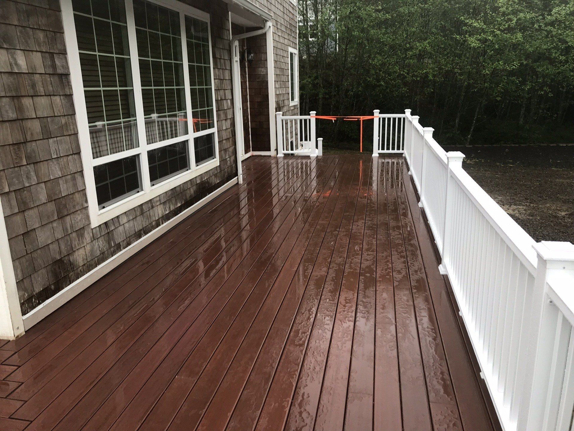Wet wooden deck with white railing next to a house with large windows.