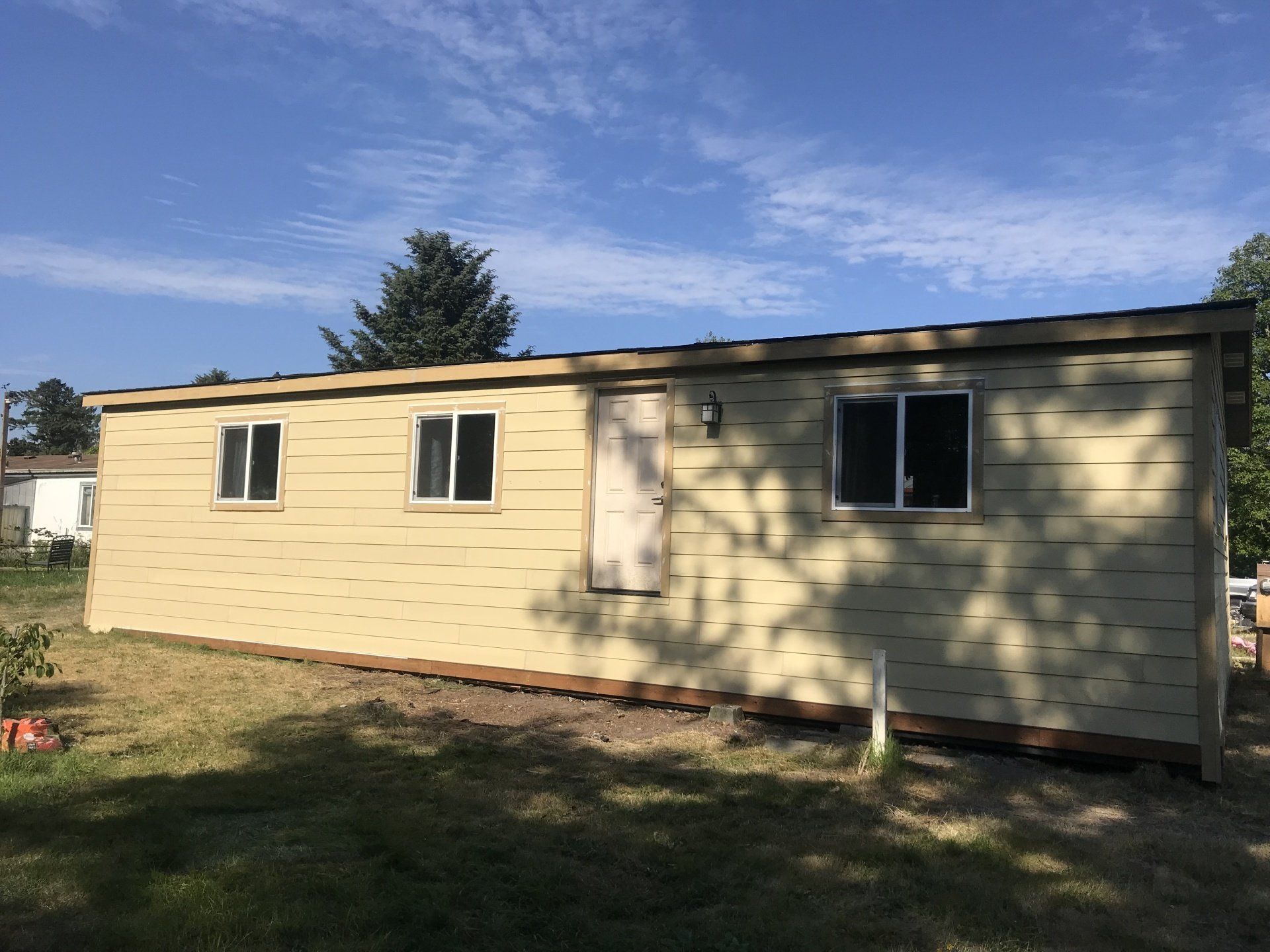 Tan shed with three windows and one door, set against a blue sky with green grass.