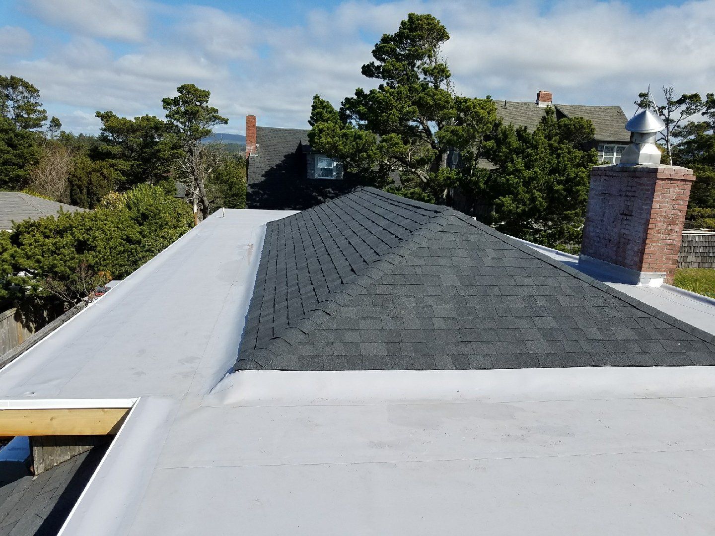 View of residential roofs: flat gray roof with an angled, shingled roof and a brick chimney under a cloudy blue sky.
