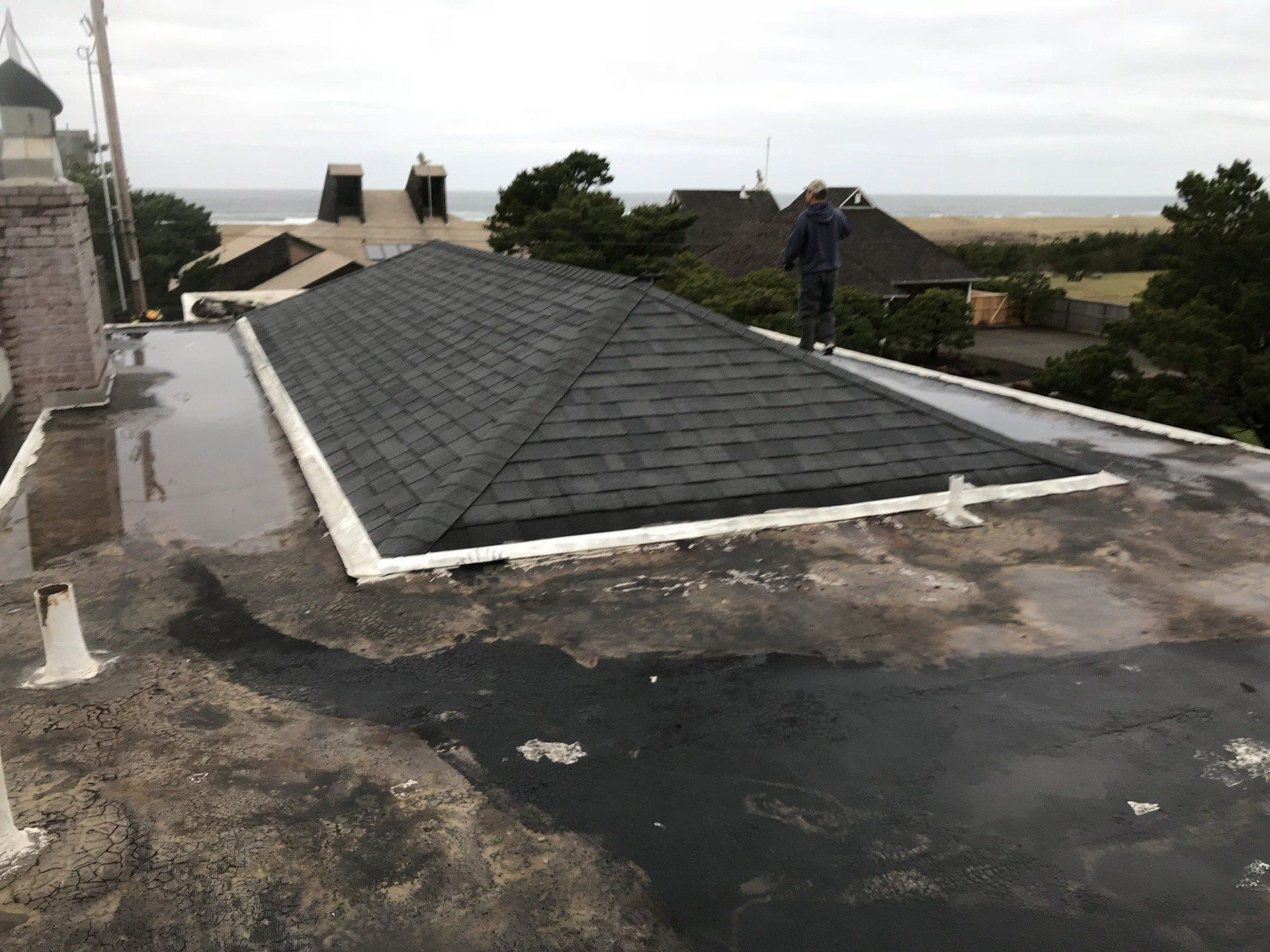 A person stands on a flat roof next to a dark shingle roof, overlooking a beach.