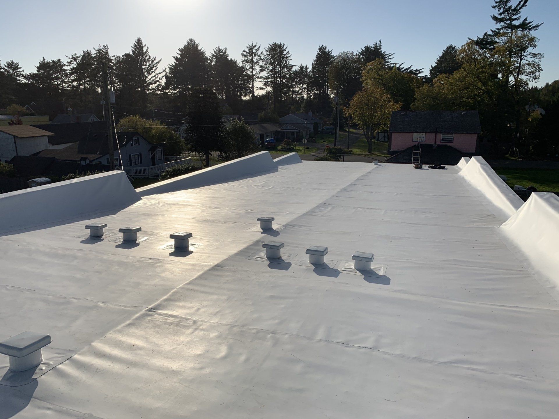 White flat roof with vents, trees, and buildings in the background under a sunny sky.