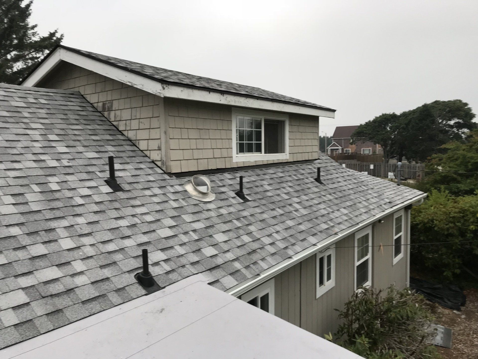 Gray asphalt shingle roof with vents, a dormer, and white siding on a house. Overcast sky.