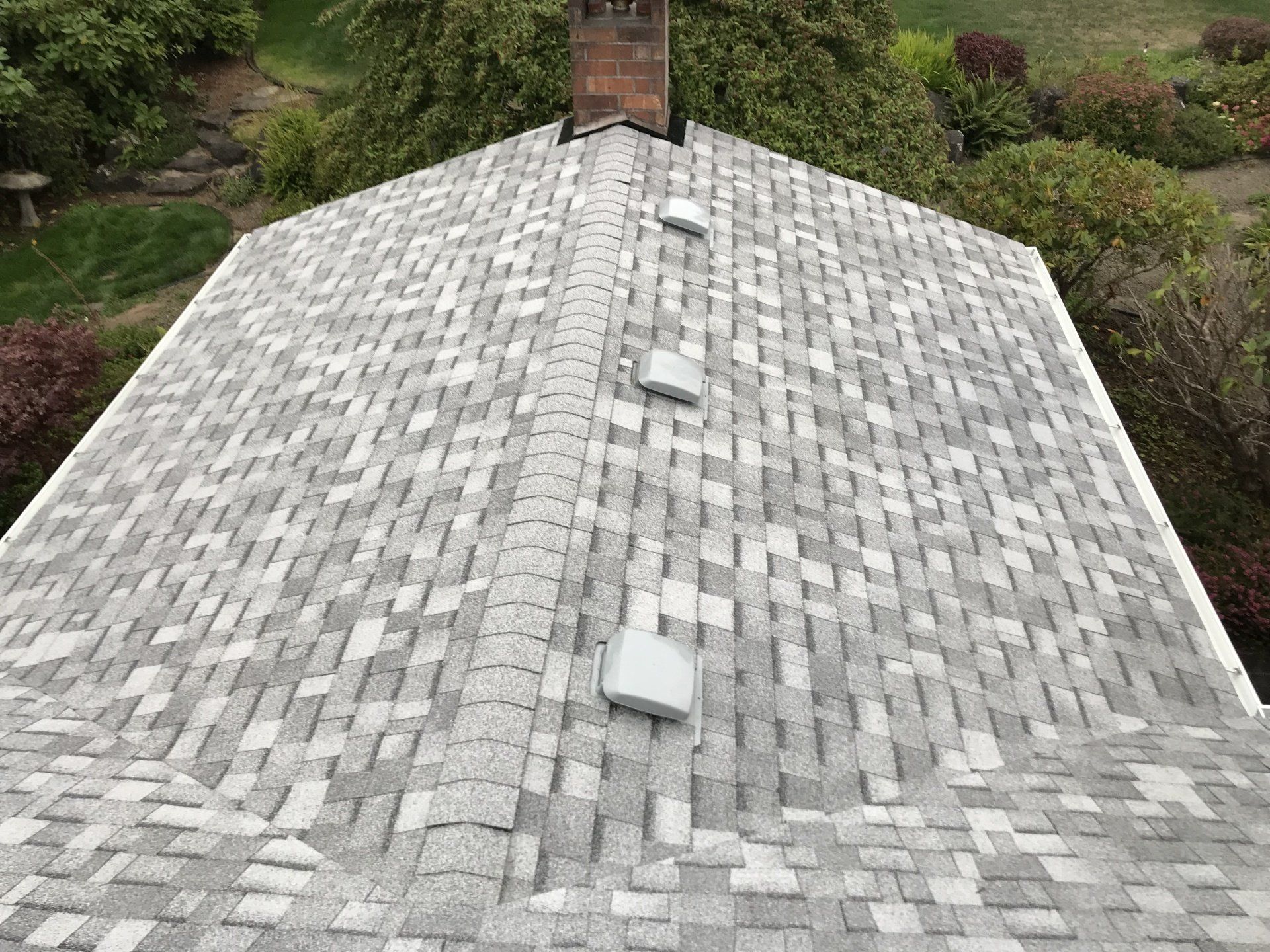 Gray and white asphalt shingle roof with three square vents and a brick chimney against a backdrop of greenery.