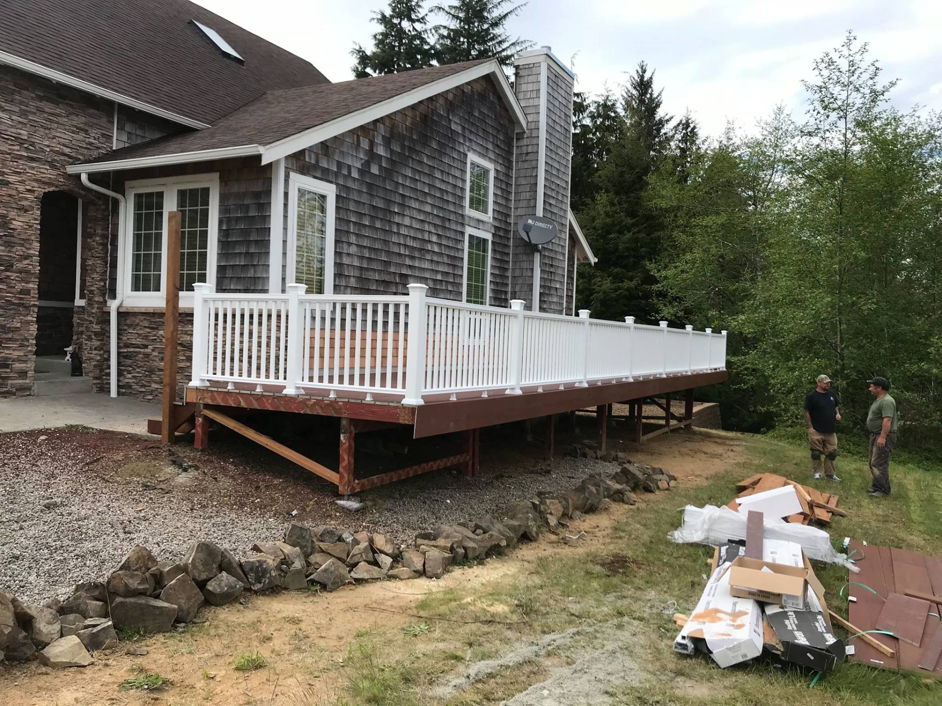 A house with a white-railed deck under construction. Two men stand nearby, with building materials scattered.