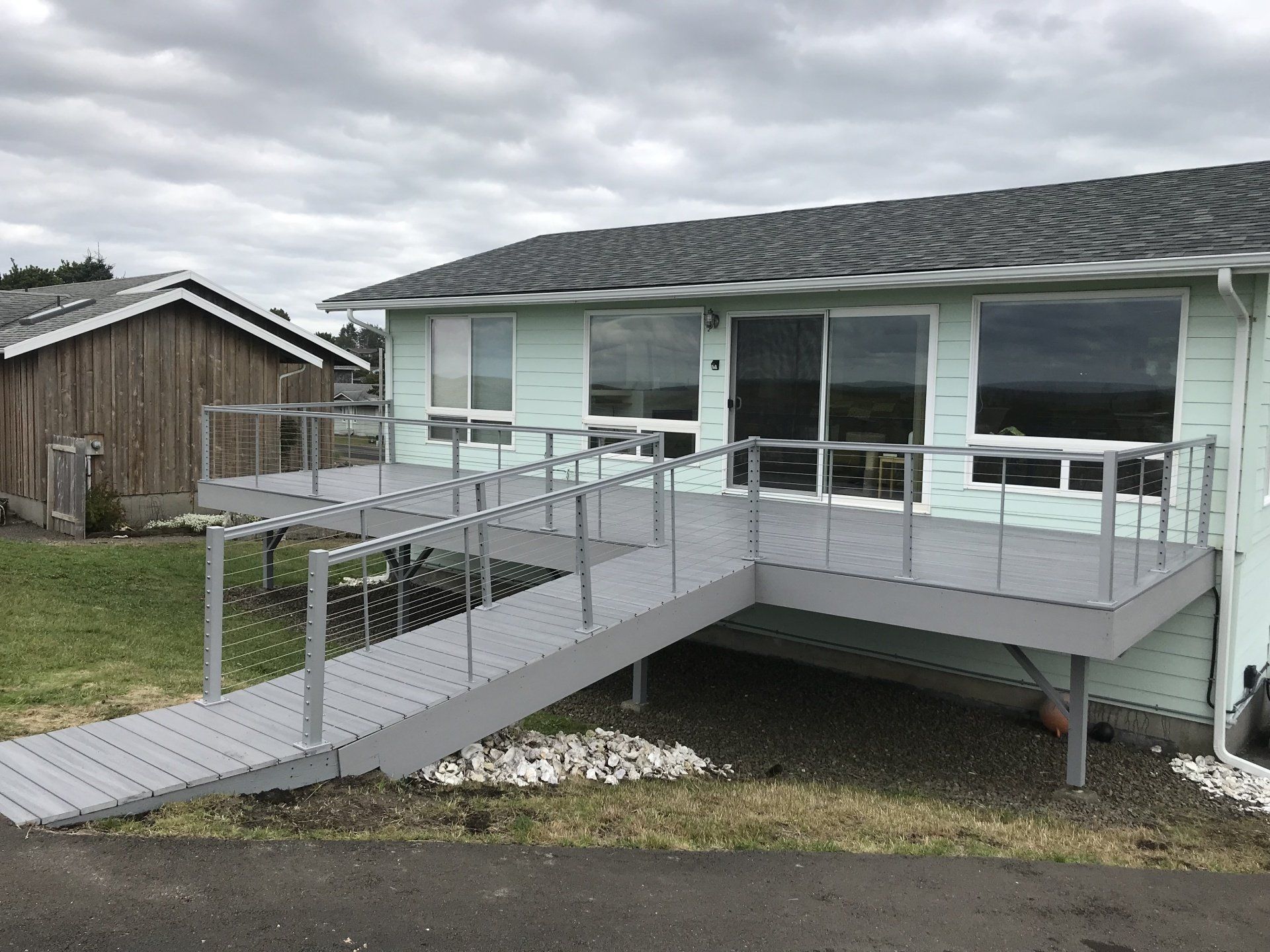 A house with a gray deck and ramp. Wooden shed in the background. Overcast sky.