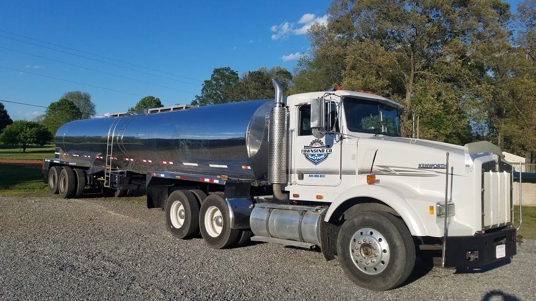 A white semi truck with a tanker trailer attached to it is parked in a gravel lot.