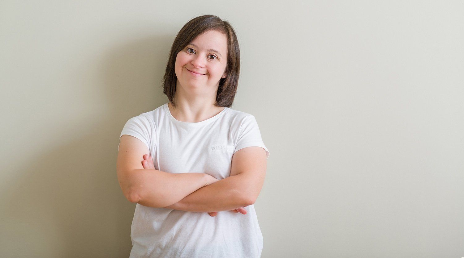 A woman with down syndrome is standing with her arms crossed and smiling.