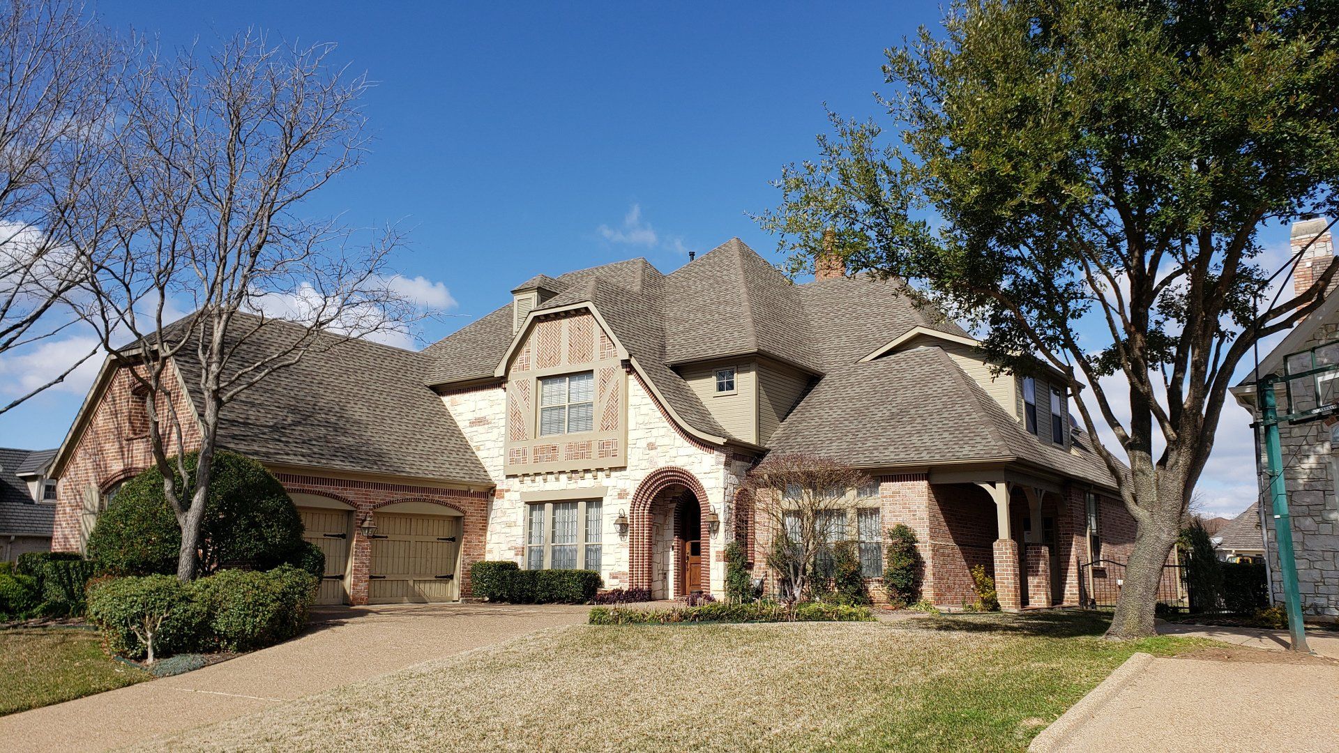 Large tan brick suburban house with a steep roof, arched entry, and leafless trees in front