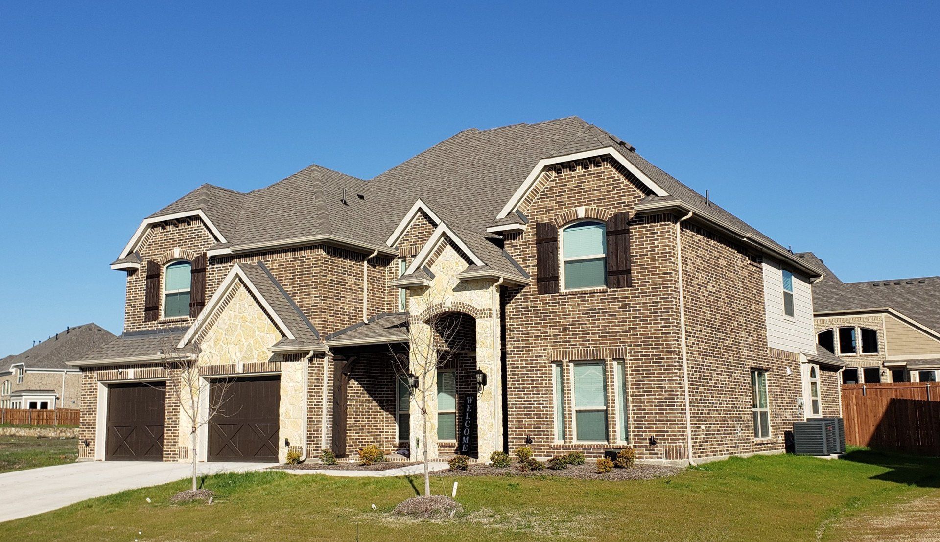 Large two-story brick house with a tiled roof and attached garage on a sunny suburban lot