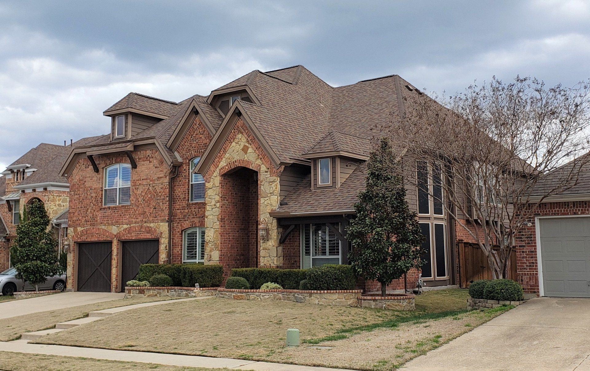 Large brick suburban house with a brown roof, arched entry, and attached garage in a front yard