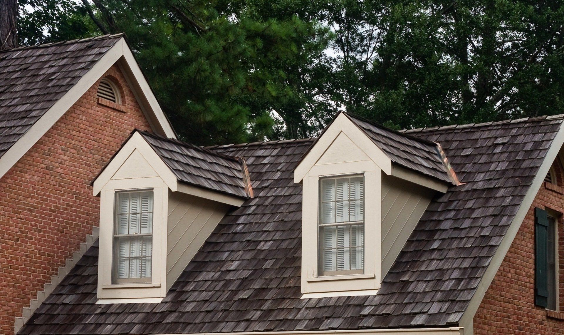 Two dormer windows on a shingled roof beside a red brick house and trees