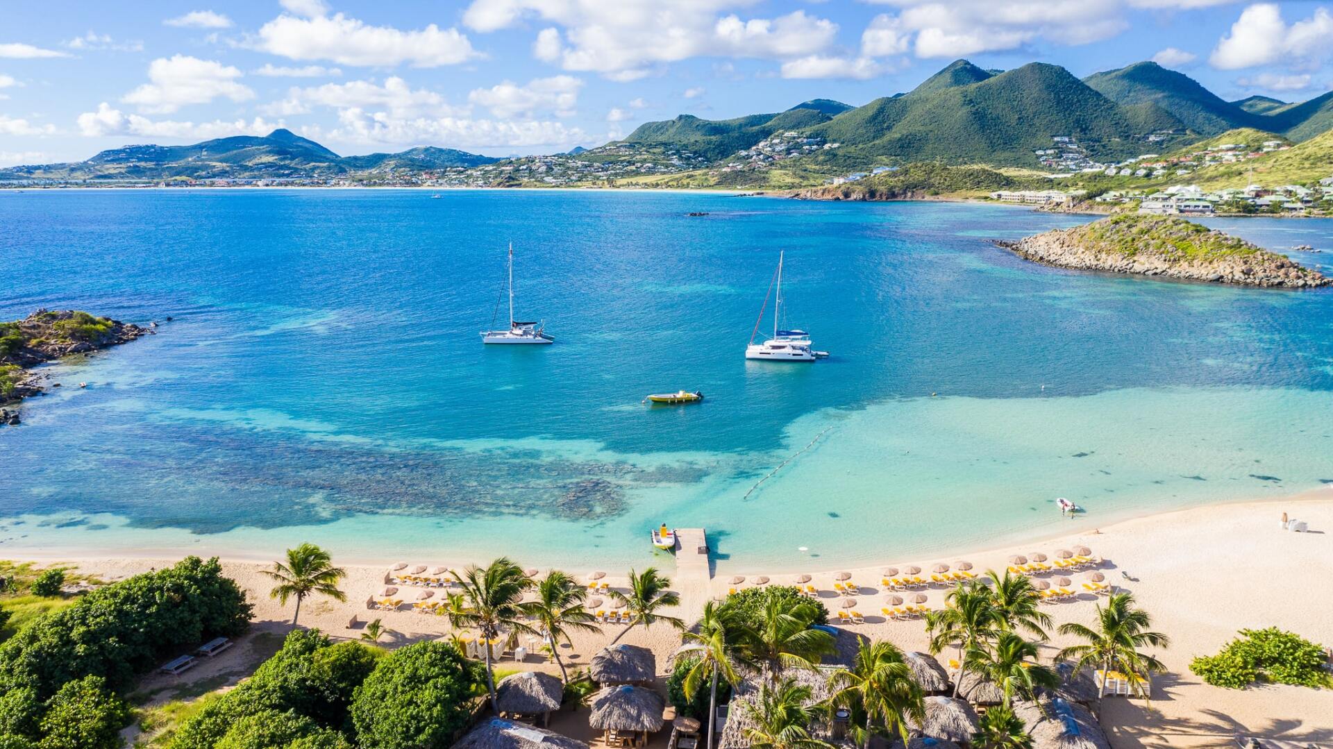 An aerial view of a tropical beach with boats in the water and mountains in the background.