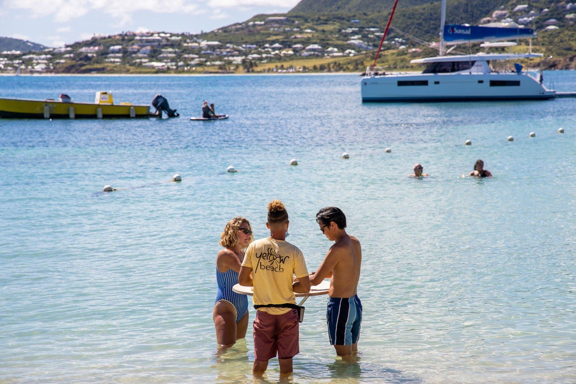 A group of people are standing in the water on a beach with a boat in the background.