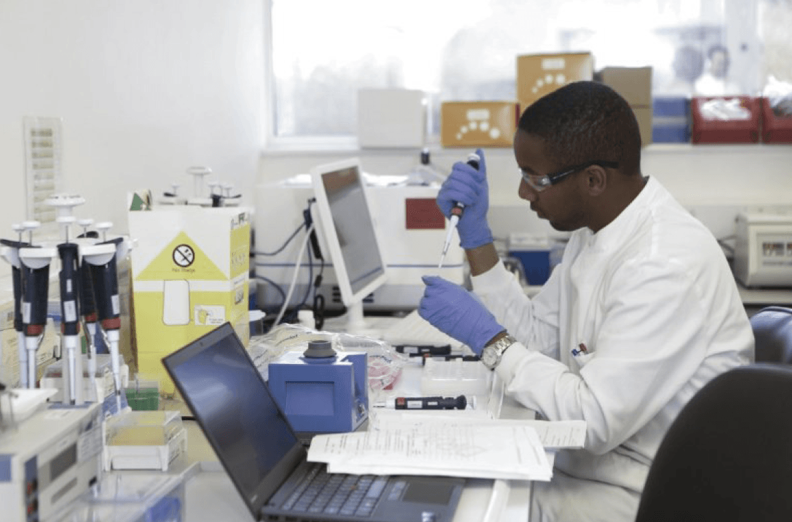 A man does labwork at a desk with a computer, holding a test tube and pipette with gloves, from Genentech Campus Plan 2019 PDF.