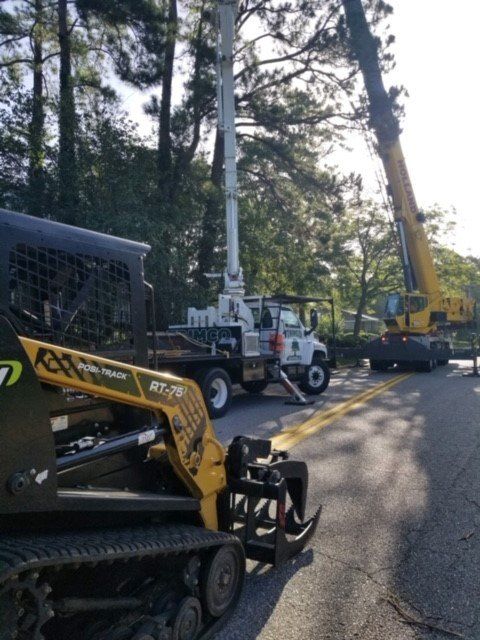 A yellow tracked skid steer and a tree-trimming truck on a road. A crane is in the background.
