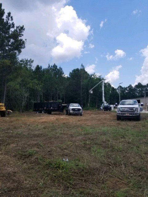 Construction site with trucks, a tree, and a lift against a blue sky with clouds.