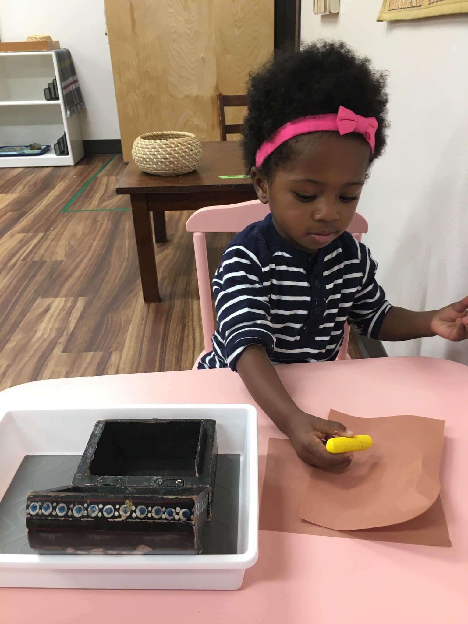 A little girl is sitting at a table holding a yellow crayon.