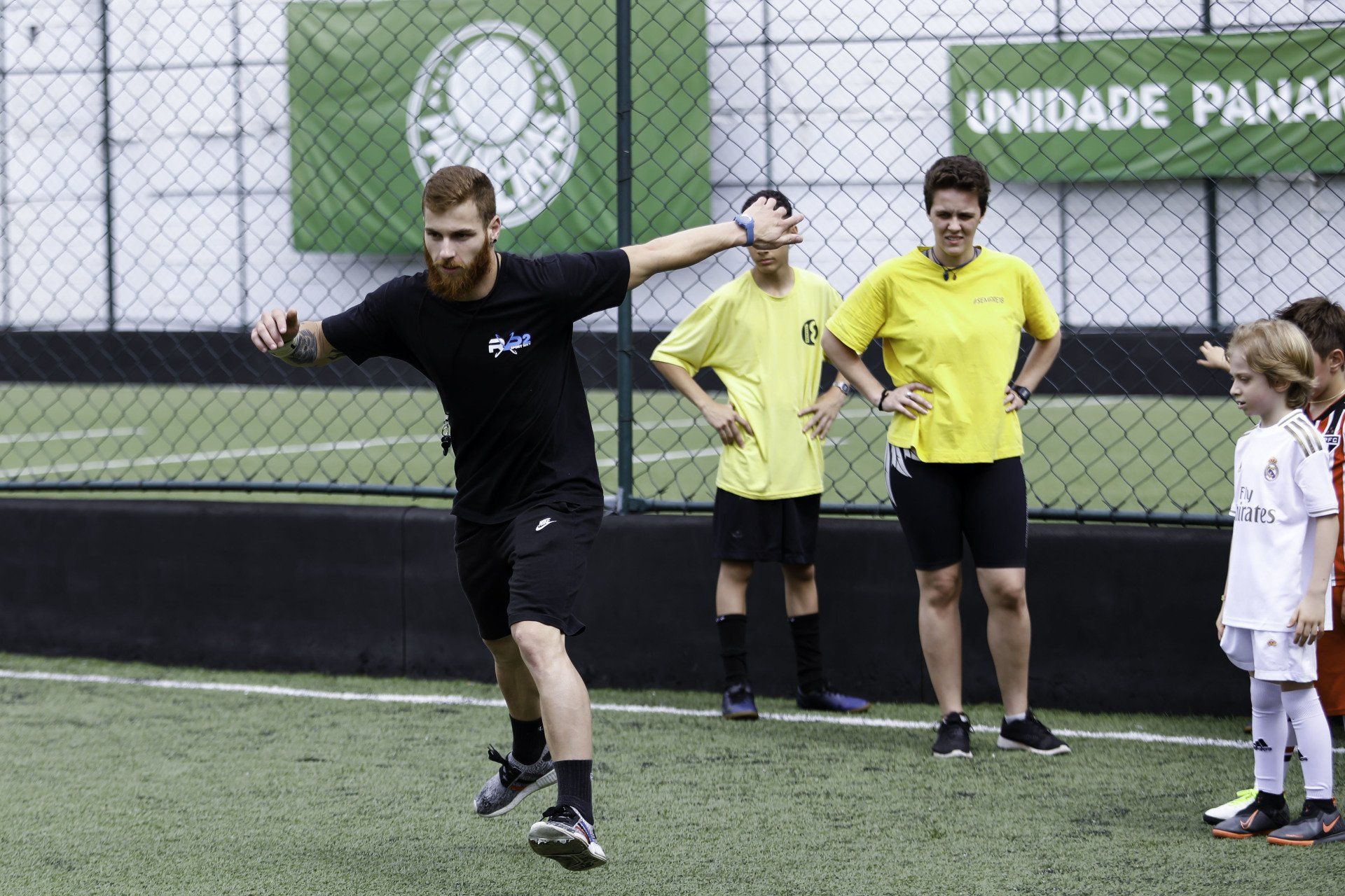 Um homem vestido de preto demonstra um lance de futebol em um campo verde enquanto crianças e um árbitro observam.
