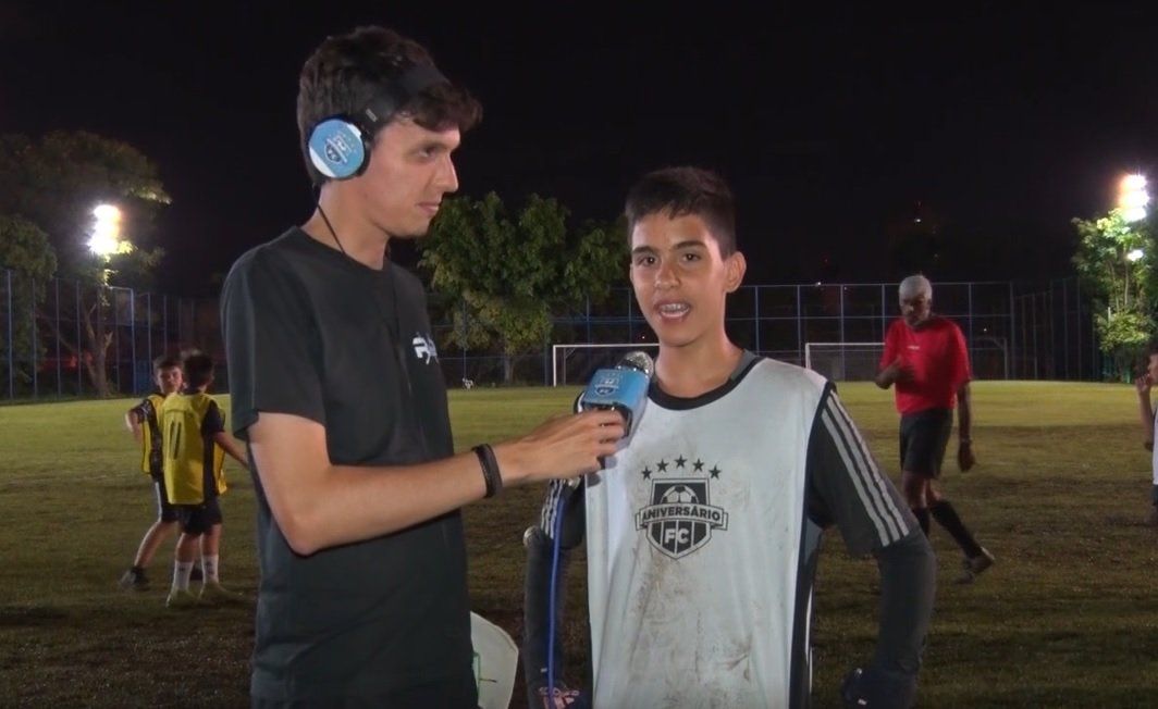 Homem entrevistando um menino vestindo um colete de treino de futebol em um campo iluminado à noite.