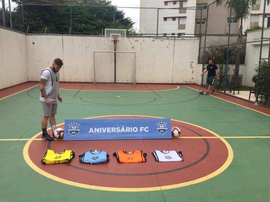 Dois homens em uma quadra de futsal com bolas de futebol, coletes de time e uma faixa com os dizeres