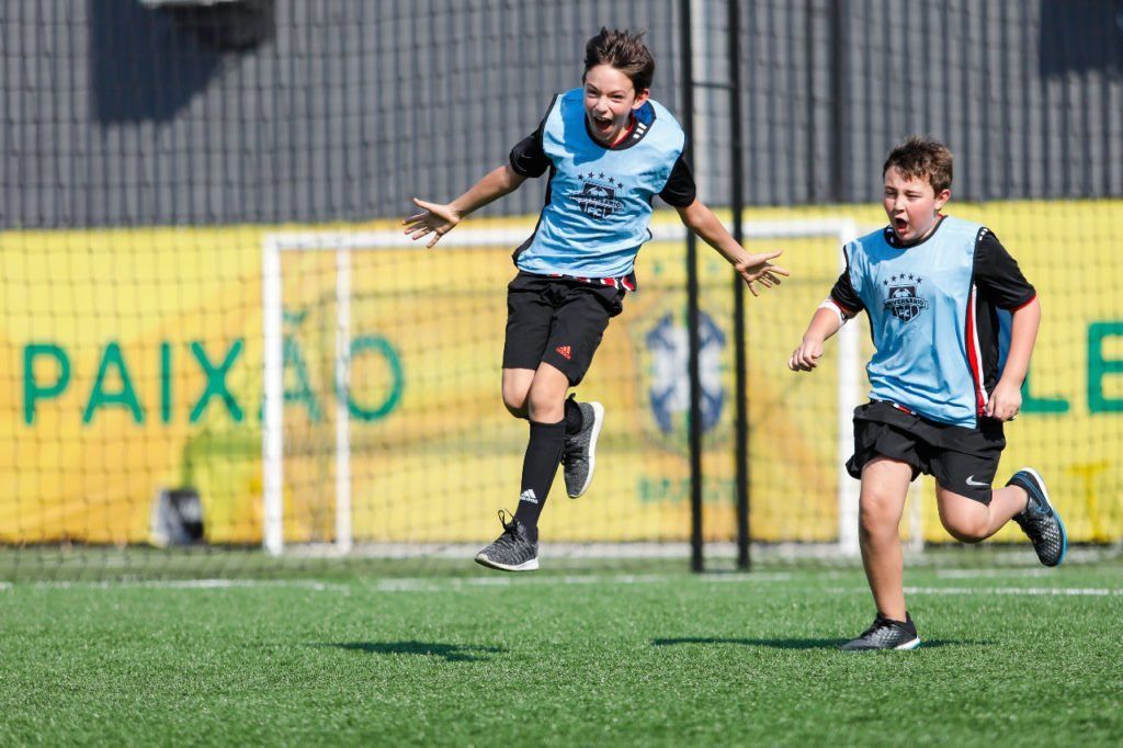 Dois meninos comemorando em um campo de futebol. Um pula no ar, sorrindo, enquanto o outro corre ao lado. Campo verde, faixa amarela.