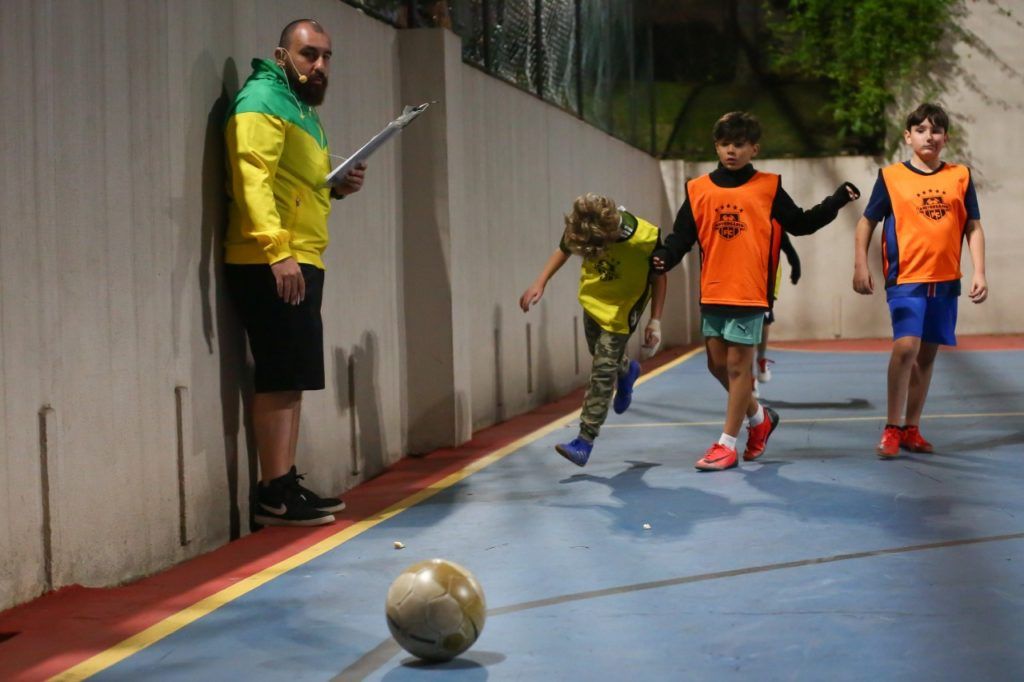 O treinador de futebol observa as crianças jogando em uma quadra. Os jogadores usam coletes; a bola rola.