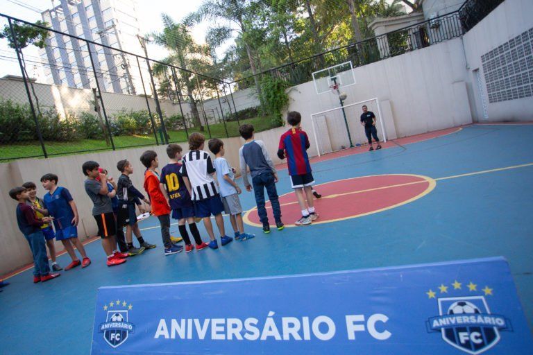 Crianças faziam fila para chutar uma bola de futebol em direção a um gol em uma quadra azul; faixa