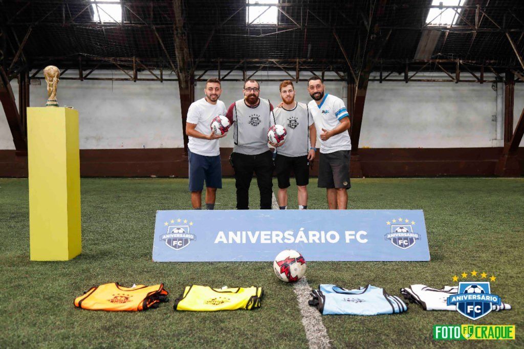 Quatro homens com bolas e camisas de futebol posam com um troféu e uma faixa em um campo verde.