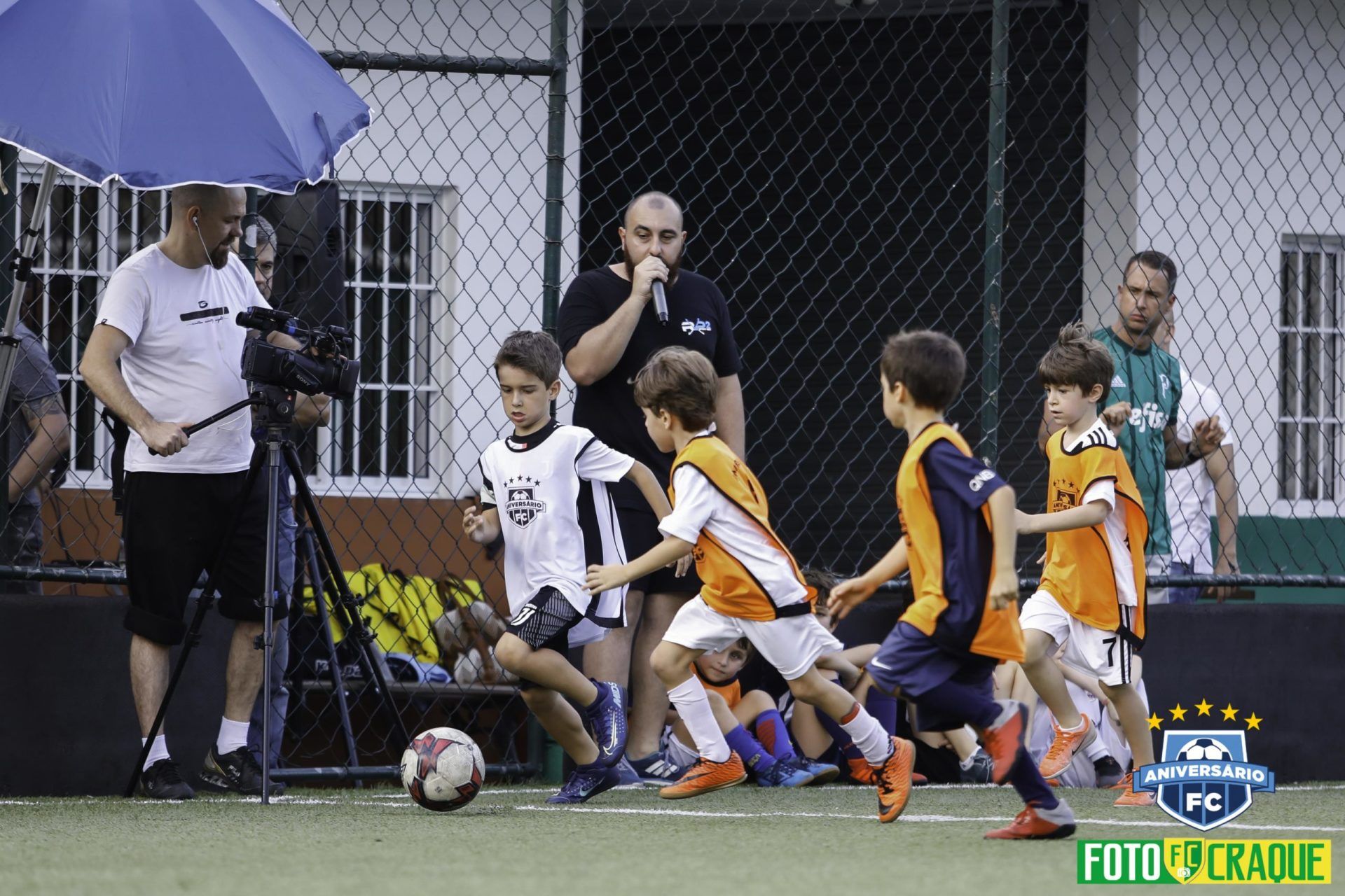 Crianças jogando futebol em um campo enquanto um adulto filma. Alguns jogadores vestem camisas laranja e branca.